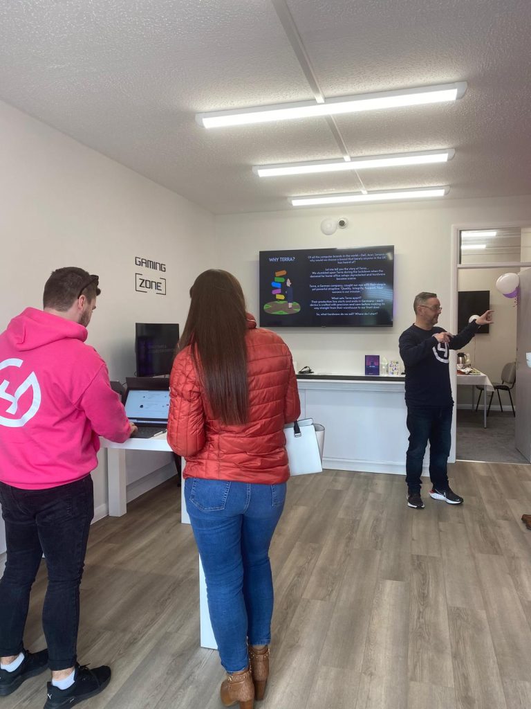 The image depicts a lively scene inside a tech store or event space. In the foreground, a person in a pink hoodie is interacting with a laptop, and a woman in a red jacket is standing nearby, engaged in the activity. In the background, another individual in a dark shirt is speaking or demonstrating something, with a large monitor displaying "Why Terra?" on the wall. The room has clean, modern decor with bright lighting and a designated "Gaming Zone" area visible on the left. The space exudes a welcoming and community-focused atmosphere, ideal for tech events or workshops