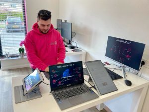 The image shows a person wearing a bright pink hoodie, standing at a desk with multiple electronic devices, including laptops, a tablet, and monitors. The workspace appears to be a clean and modern tech environment with natural light coming through large windows in the background. The monitors display product information, including a "Jabra Headset" and "Terra All-In-One PC." The scene suggests a tech demonstration or setup session.